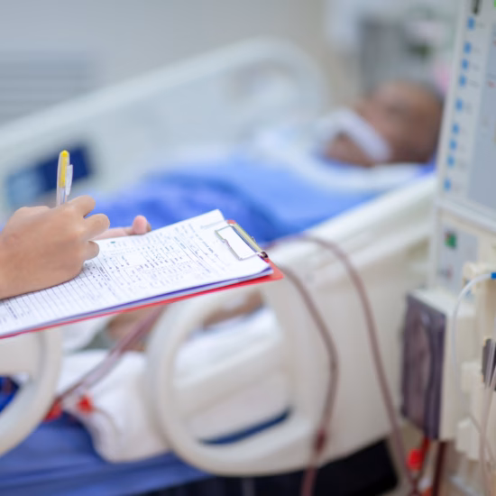 Ziekenhuisomgeving met medisch apparaat naast een patiëntbed en een zorgmedewerker die gegevens noteert op een formulier.