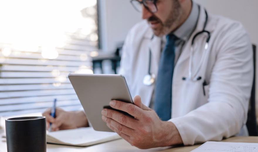 Zorgprofessional met stethoscoop gebruikt een tablet en maakt aantekeningen aan een bureau in een medische werkomgeving.