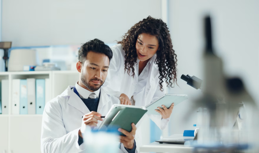 Science, talking and people on tablet in lab with microscope for analysis, medical research and study. Biology, pharmaceutical team and man with woman on tech for results, review and online report.