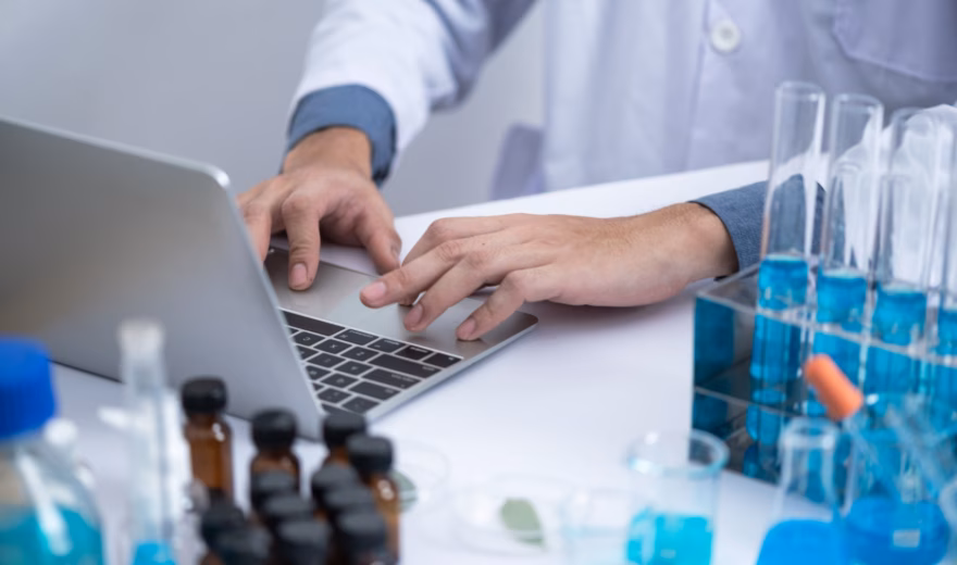 Ein Laborassistent führt neben Reagenzgläsern und Laborproben Datenverarbeitungsaufgaben auf einem Laptop durch