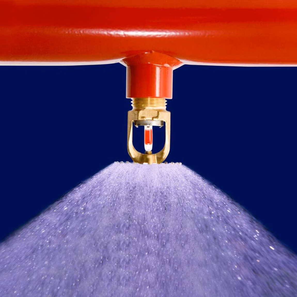 Nahaufnahme eines Sprinklerkopfes an einer roten Leitung, der einen gleichmäßigen Wassernebel versprüht. Das Bild veranschaulicht ein automatisches Löschsystem für den Brandschutz in Gebäuden und industriellen Umgebungen.