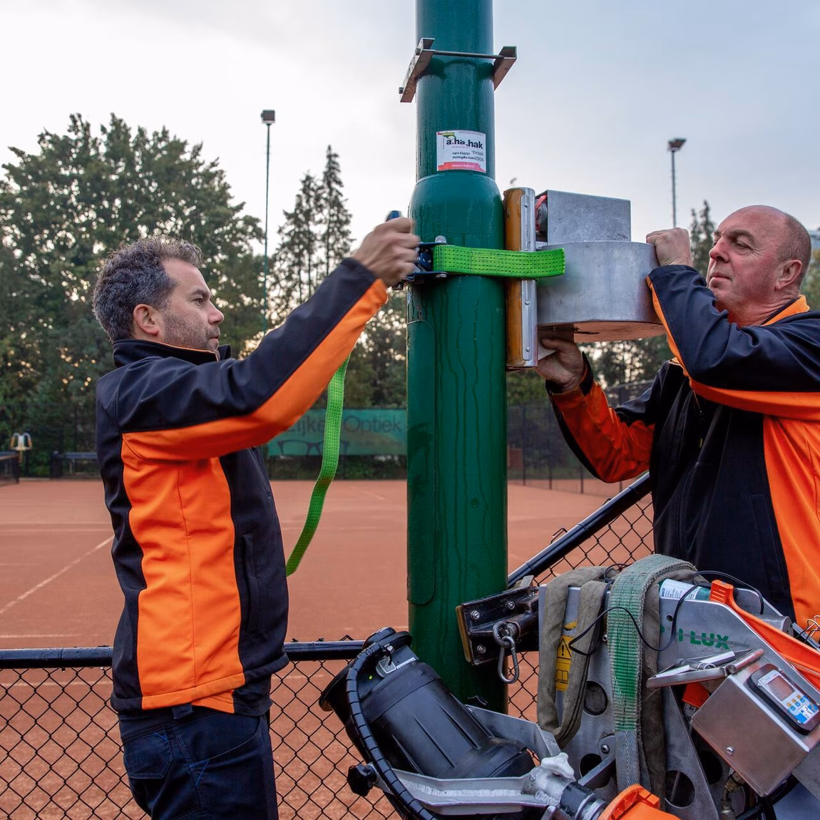 Des employés inspectent un mât d'éclairage sur un terrain de sport à l'aide d'une nacelle élévatrice.