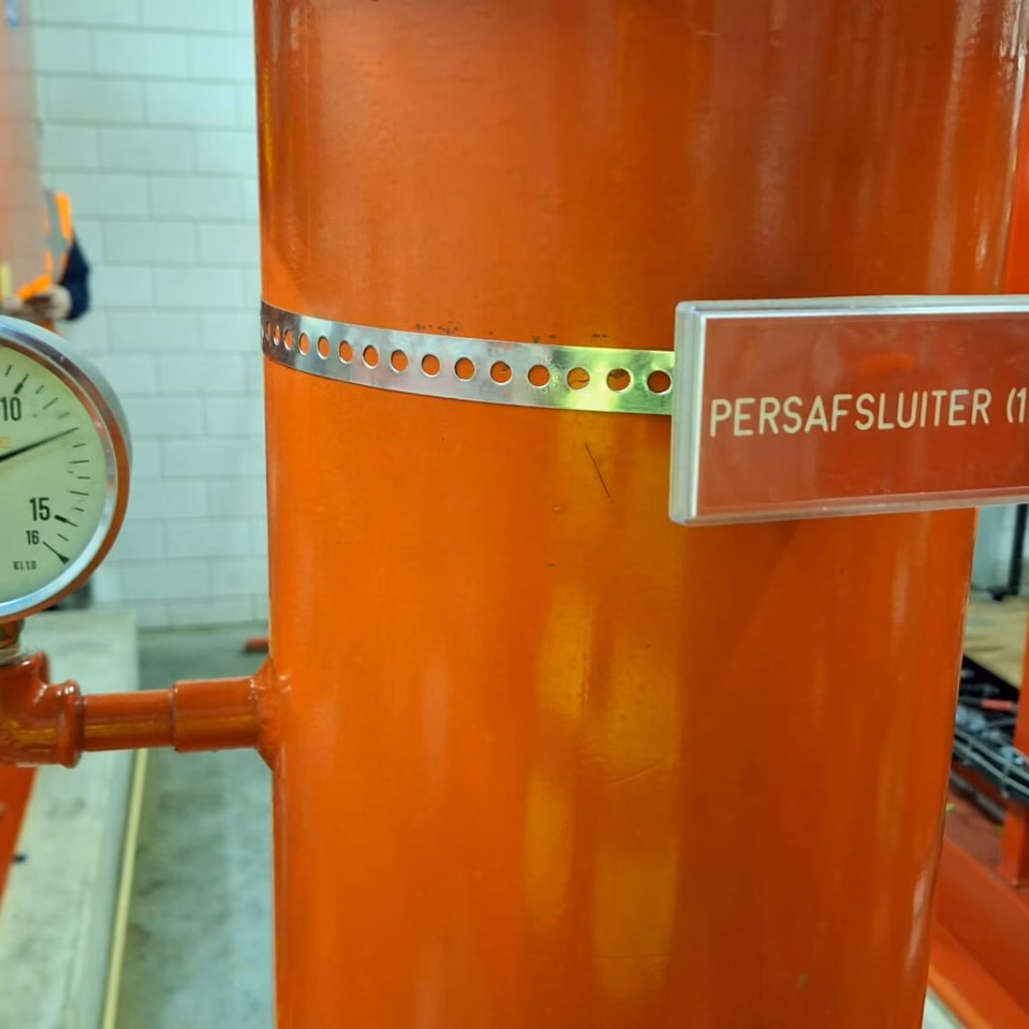 Persafsluiter op een oranje leidinginstallatie met manometer in een technische ruimte.
