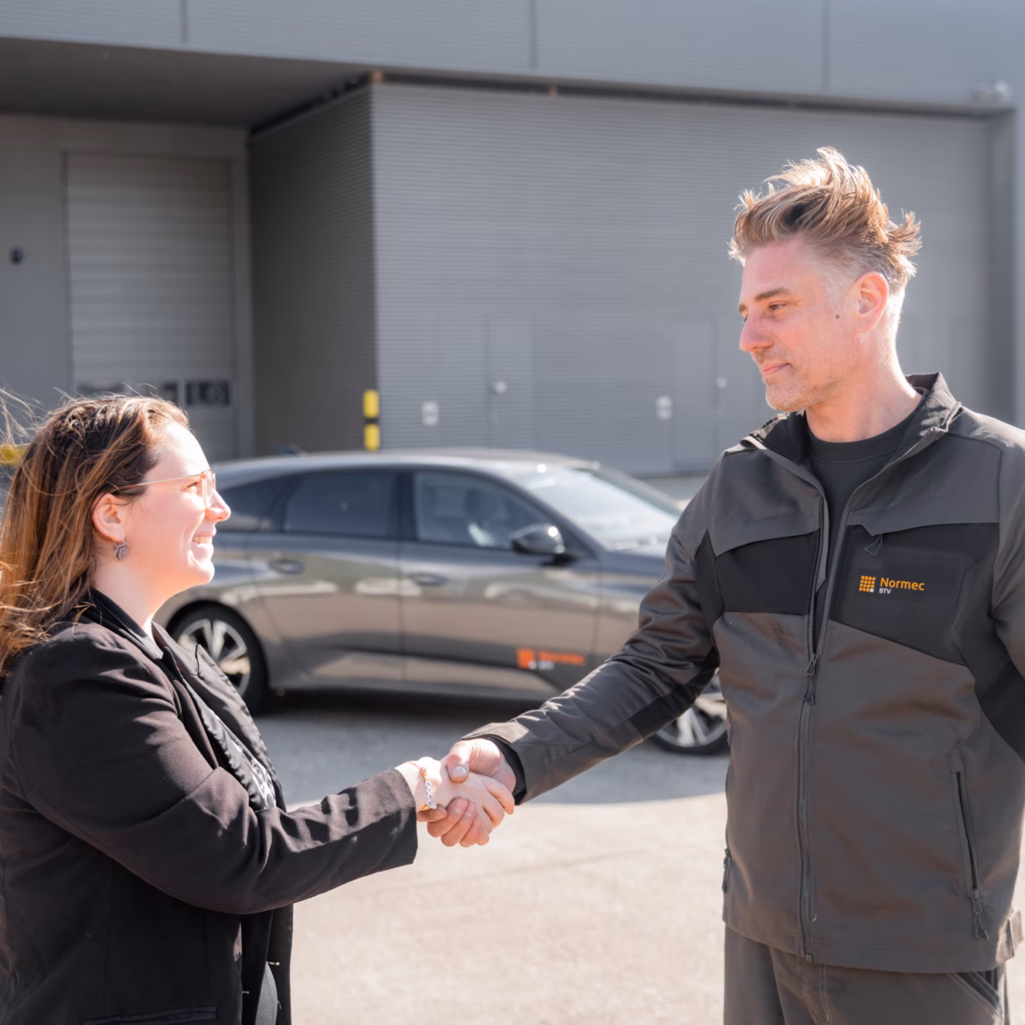 Op een buitenterrein voor een bedrijfspand schudden twee personen elkaar de hand. Op de achtergrond zijn een geparkeerde auto en een bedrijfsgebouw zichtbaar. De afbeelding straalt samenwerking, klantcontact en professionele dienstverlening uit.