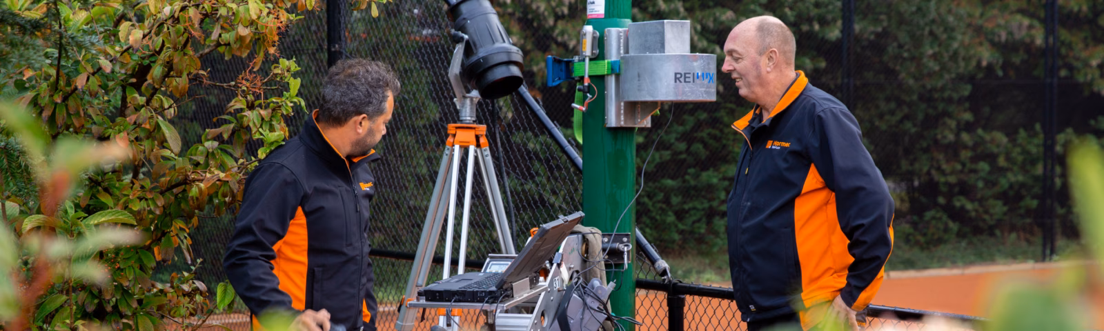Two technicians are conducting surveying work with a total station next to a light pole at a sports facility.