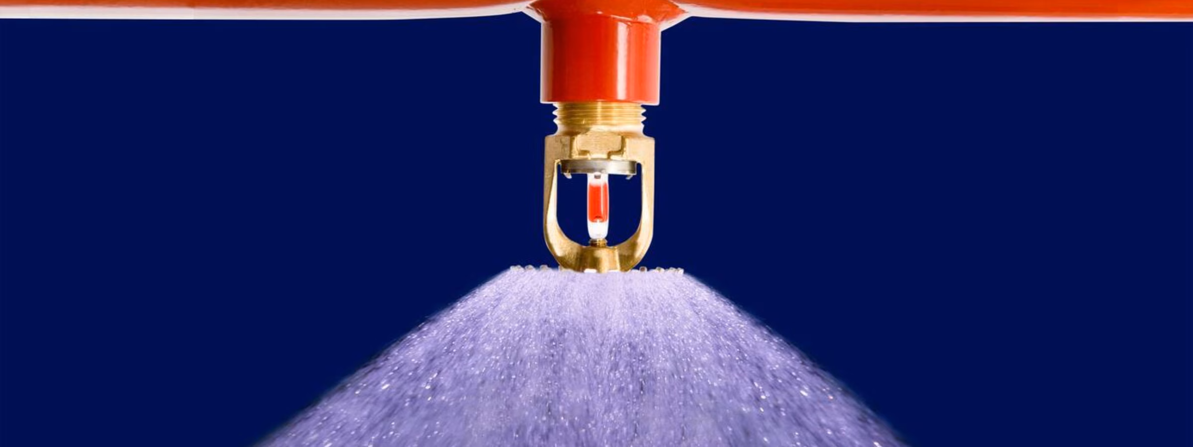 Nahaufnahme eines Sprinklerkopfes an einer roten Leitung, der einen gleichmäßigen Wassernebel versprüht. Das Bild veranschaulicht ein automatisches Löschsystem für den Brandschutz in Gebäuden und industriellen Umgebungen.