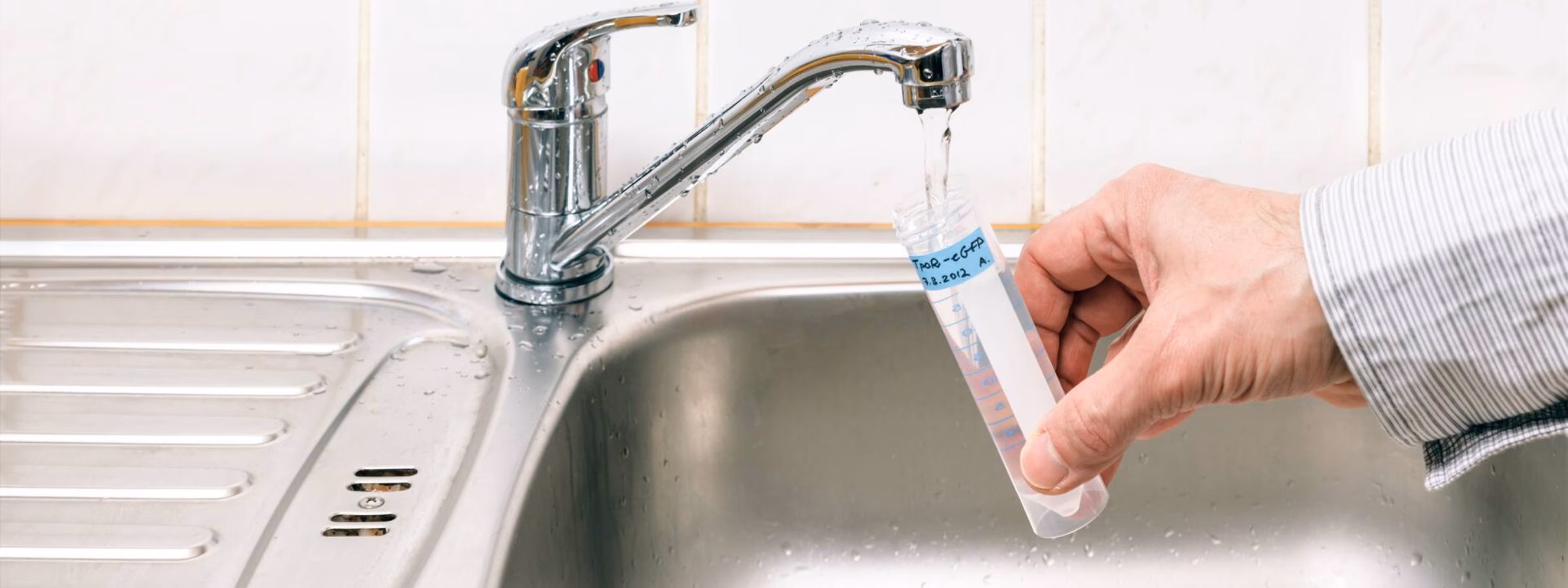 A water sample is collected from a kitchen faucet above a stainless steel sink.