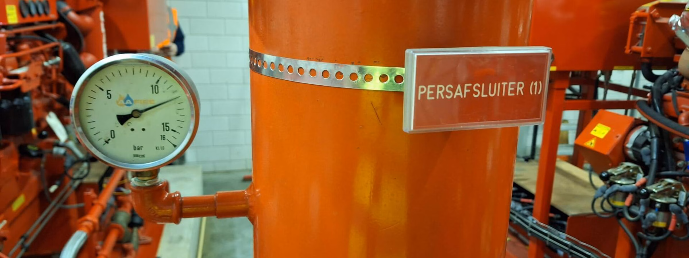 Persafsluiter op een oranje leidinginstallatie met manometer in een technische ruimte.