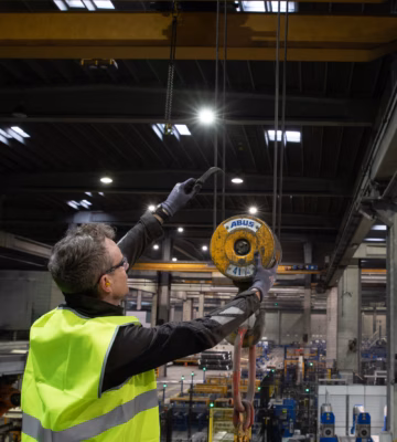 Industrial inspector wearing high-visibility safety clothing conducts an inspection of an overhead lifting hoist in a manufacturing facility, highlighting crane inspections, lifting equipment certification, workplace safety, regulatory compliance, preventive maintenance, and quality assurance for industrial lifting operations.