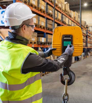 Industrial inspector wearing high-visibility safety clothing uses a handheld control device to operate and monitor a suspended lifting hook in a warehouse environment, highlighting load testing, lifting equipment inspections, safety compliance, equipment certification, and quality assurance for material handling operations.