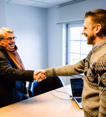 Twee personen begroeten elkaar met een handdruk aan een vergadertafel in een kantooromgeving.