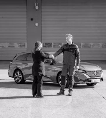 Twee personen ontmoeten elkaar op een bedrijfsterrein naast een geparkeerde auto.