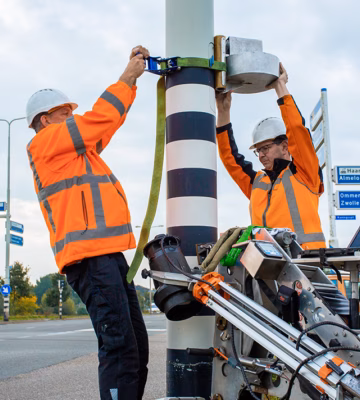 Twee medewerkers controleren apparatuur bij een verkeerspaal langs de weg.