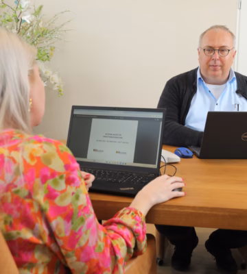 Twee personen zitten tegenover elkaar aan een tafel en werken met laptops tijdens een gesprek.