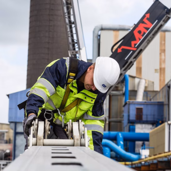 Medewerker in veiligheidskleding inspecteert een ankerpunt en valbeveiligingssysteem op hoogte bij een industriële installatie.