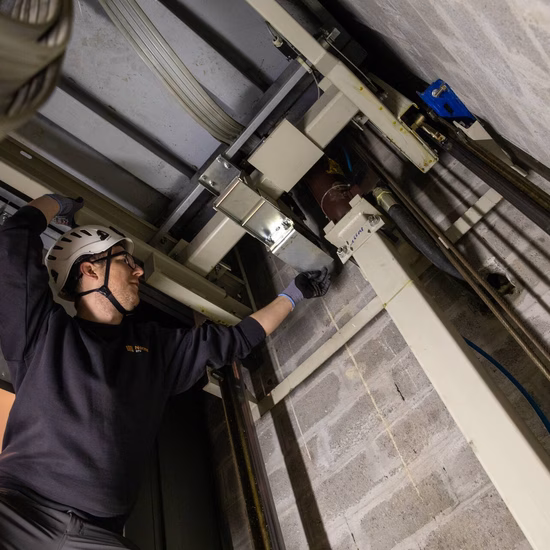 Technicus voert onderhoud uit aan een liftinstallatie in een technische ruimte met zichtbare bekabeling en mechanische onderdelen.
