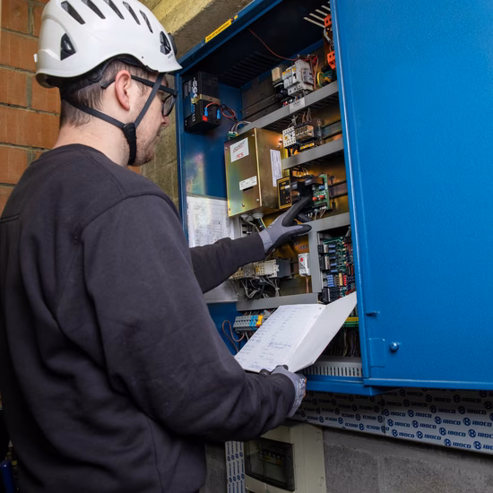 Technicus met veiligheidshelm controleert een elektrische installatie in een schakelkast en gebruikt een tablet voor registratie.