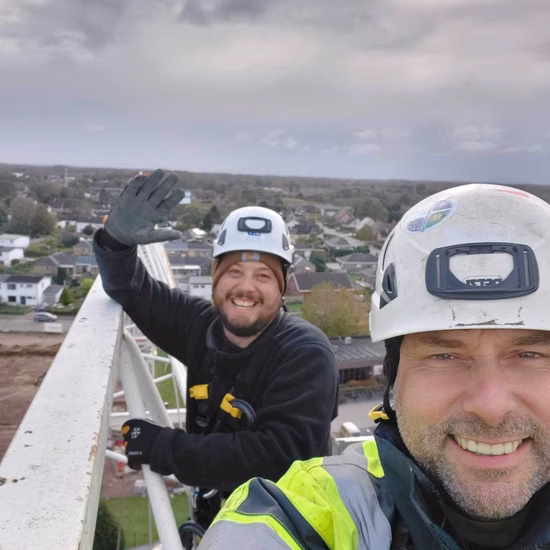 Twee monteurs voeren werkzaamheden uit op hoogte aan een gevelinstallatie en dragen veiligheidshelmen en persoonlijke beschermingsmiddelen.