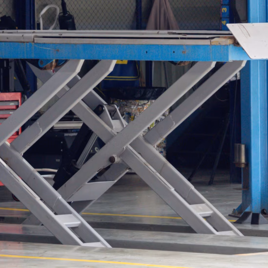 Industrial lifting platform in workshop, metal legs extended. Blue frame supports heavy machinery above. Floor marked with yellow safety lines. Tools and equipment visible in background