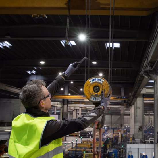 Industrial inspector wearing high-visibility safety clothing conducts an inspection of an overhead lifting hoist in a manufacturing facility, highlighting crane inspections, lifting equipment certification, workplace safety, regulatory compliance, preventive maintenance, and quality assurance for industrial lifting operations.