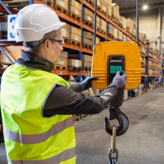 Industrial inspector wearing high-visibility safety clothing uses a handheld control device to operate and monitor a suspended lifting hook in a warehouse environment, highlighting load testing, lifting equipment inspections, safety compliance, equipment certification, and quality assurance for material handling operations.