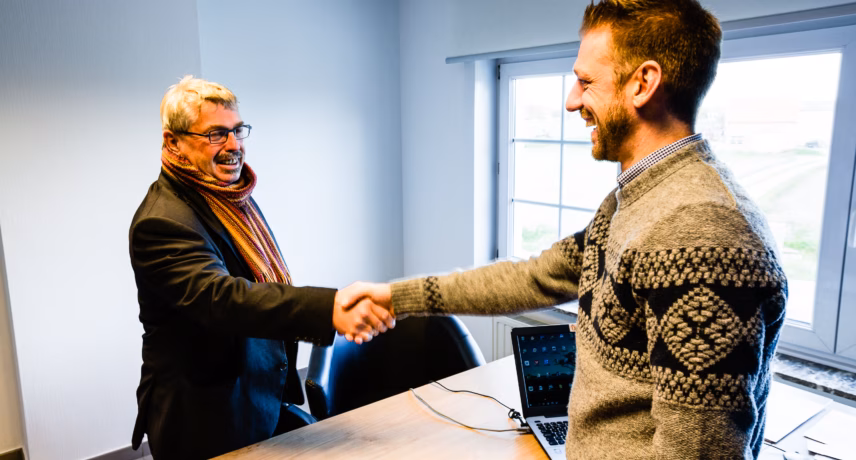Twee personen begroeten elkaar met een handdruk aan een vergadertafel in een kantooromgeving.