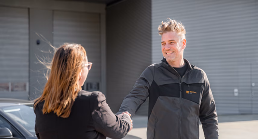 Twee personen begroeten elkaar op een bedrijfsterrein voor een bedrijfsgebouw.