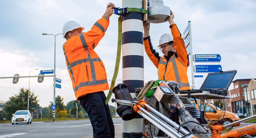Zwei Mitarbeiter überprüfen die Geräte an einem Verkehrsmast am Straßenrand.