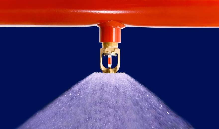 Nahaufnahme eines Sprinklerkopfes an einer roten Leitung, der einen gleichmäßigen Wassernebel versprüht. Das Bild veranschaulicht ein automatisches Löschsystem für den Brandschutz in Gebäuden und industriellen Umgebungen.