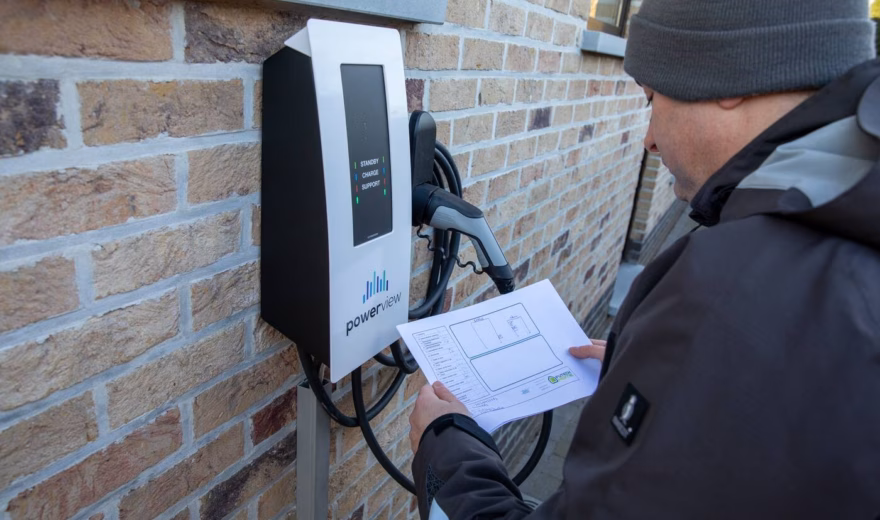 Un technicien vérifie une borne de recharge fixée à un mur en briques à l'aide d'un document lors d'une inspection.