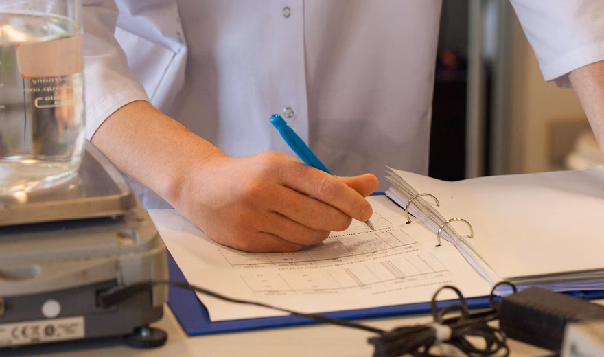 Persoon in laboratoriumjas noteert meetgegevens op een formulier naast laboratoriumapparatuur.