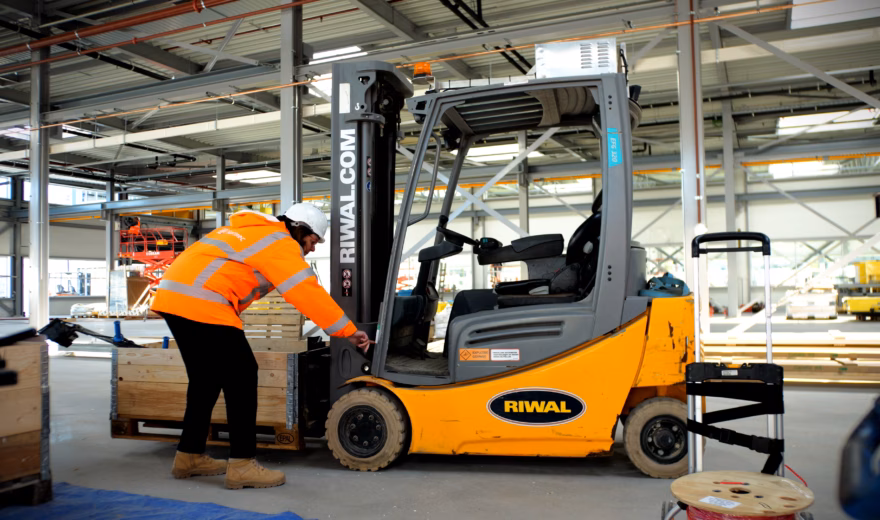 Warehouse employee wearing high-visibility safety clothing performs a pre-use inspection on a forklift in a modern industrial facility, highlighting operator training, workplace safety, equipment certification, compliance requirements, and safe material handling operations.