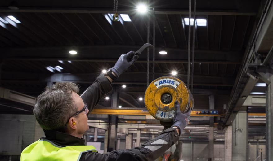 Industrial inspector wearing high-visibility safety clothing conducts an inspection of an overhead lifting hoist in a manufacturing facility, highlighting crane inspections, lifting equipment certification, workplace safety, regulatory compliance, preventive maintenance, and quality assurance for industrial lifting operations.