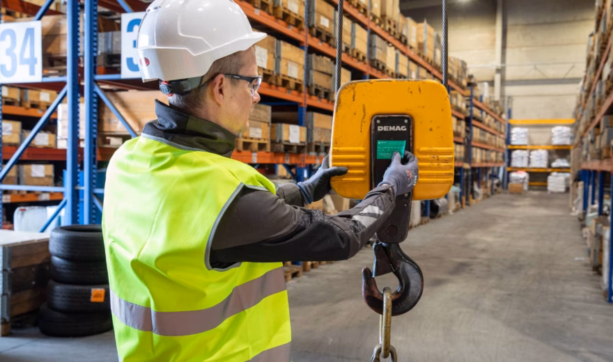 Industrial inspector wearing high-visibility safety clothing uses a handheld control device to operate and monitor a suspended lifting hook in a warehouse environment, highlighting load testing, lifting equipment inspections, safety compliance, equipment certification, and quality assurance for material handling operations.