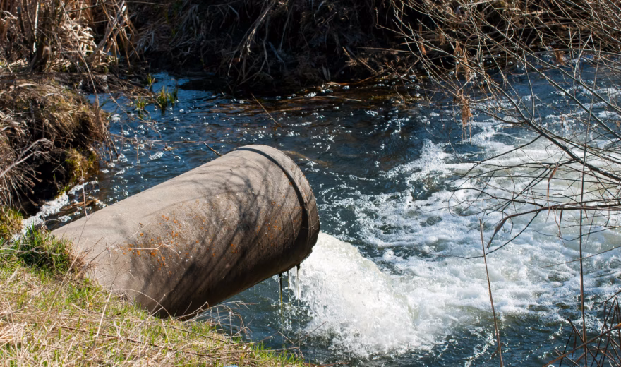 Een afvoerbuis voert water af naar een stromende beek in een natuurlijke omgeving.