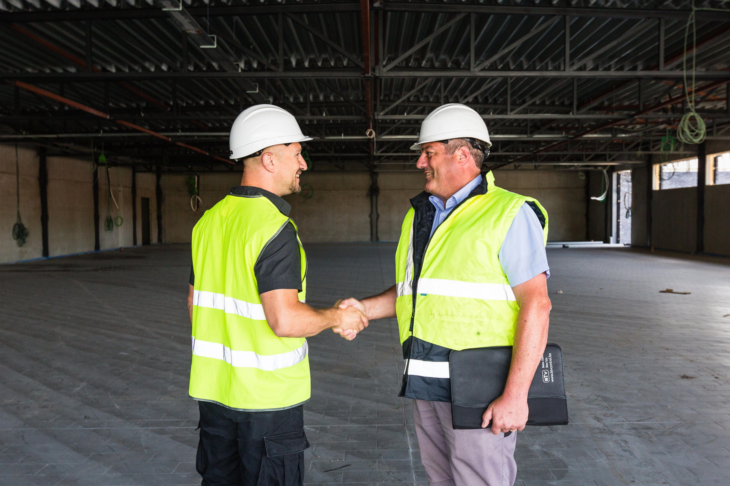 Deux personnes portant des gilets de sécurité et des casques se serrent la main dans un hangar industriel dans le cadre d'un projet de construction ou de rénovation.