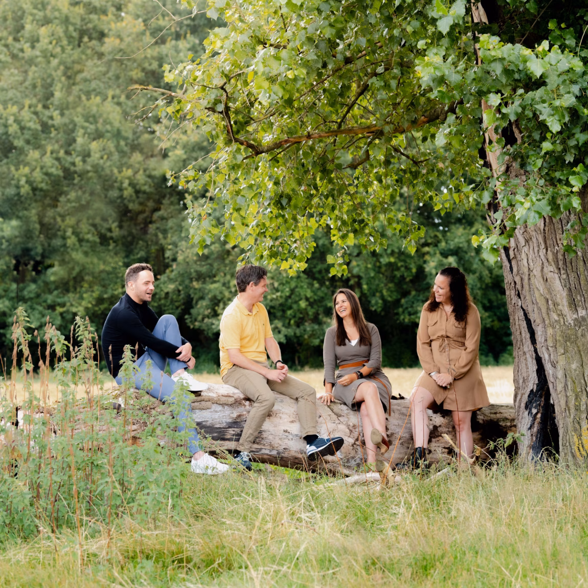 Groep mensen zit samen op boomstammen onder een grote boom in een groene natuurlijke omgeving.