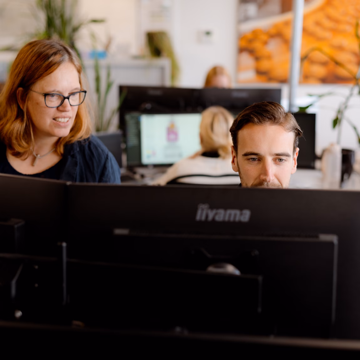 Two employees are working together behind multiple computer screens in a modern office environment.