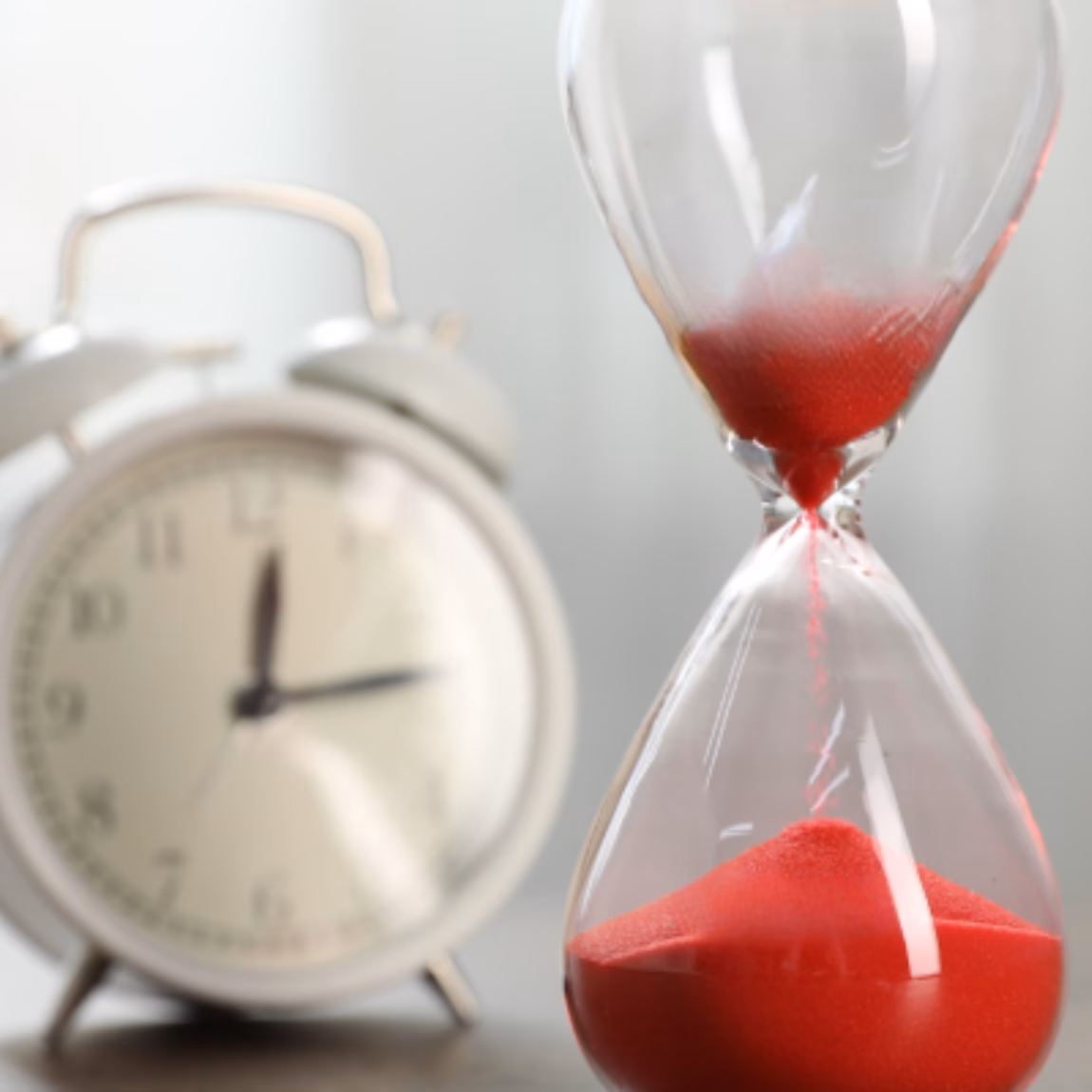 Im Vordergrund steht neben einem klassischen Wecker eine Sanduhr mit rotem Sand, die für schnelle Bearbeitungszeiten, effiziente Dienstleistungserbringung, Terminmanagement, zeitkritische Projekte und reaktionsschnellen Support bei Prüf-, Inspektions-, Zertifizierungs- und Konformitätsaktivitäten steht.