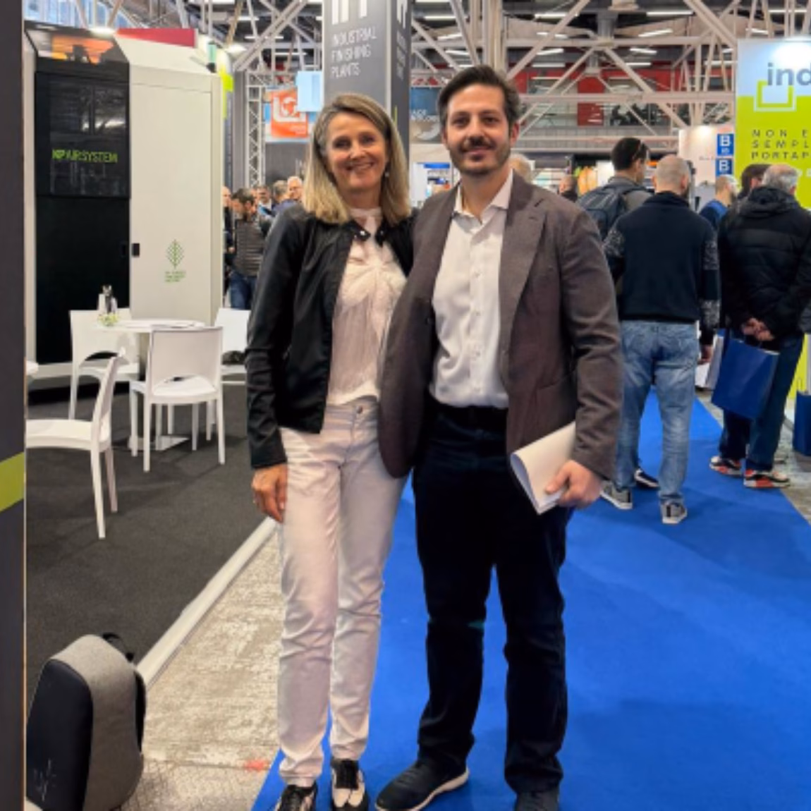 Zwei Fachleute stehen gemeinsam auf der Ausstellungsfläche einer Fachmesse, umgeben von Messeständen der Branche und Besuchern. Im Vordergrund stehen dabei geschäftliches Networking, branchenübergreifende Zusammenarbeit, Wissensaustausch sowie der Austausch über Prüf-, Inspektions-, Zertifizierungs- und Konformitätsdienstleistungen.