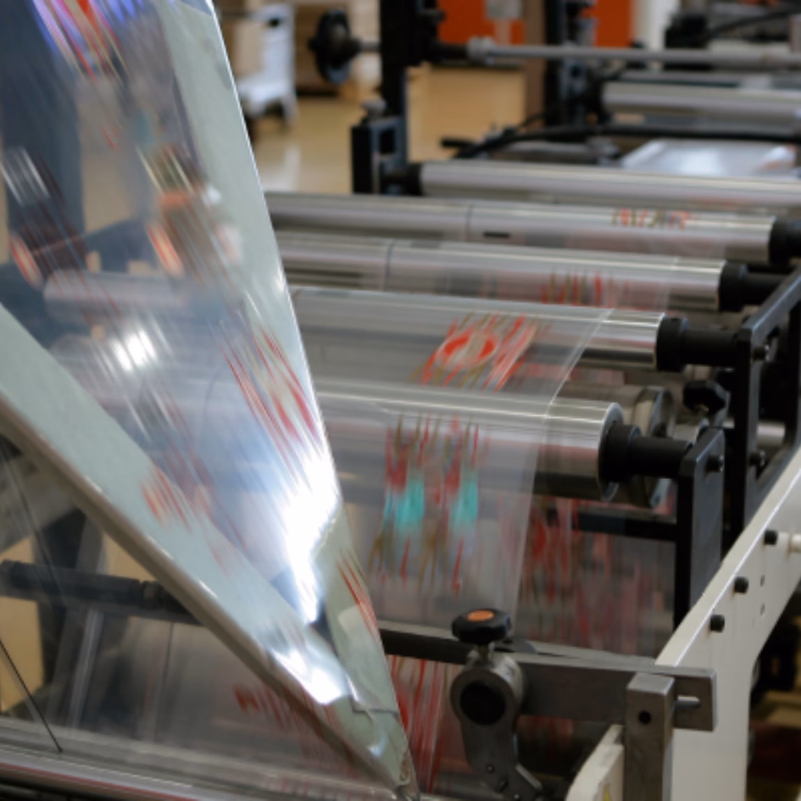 Eine transparente Verpackungsfolie durchläuft eine automatisierte Produktionslinie, die mit Walzen und Verarbeitungsmaschinen ausgestattet ist. Der Schwerpunkt liegt auf der Herstellung flexibler Verpackungen, der Qualitätskontrolle, der industriellen Fertigung und effizienten Verpackungslösungen.