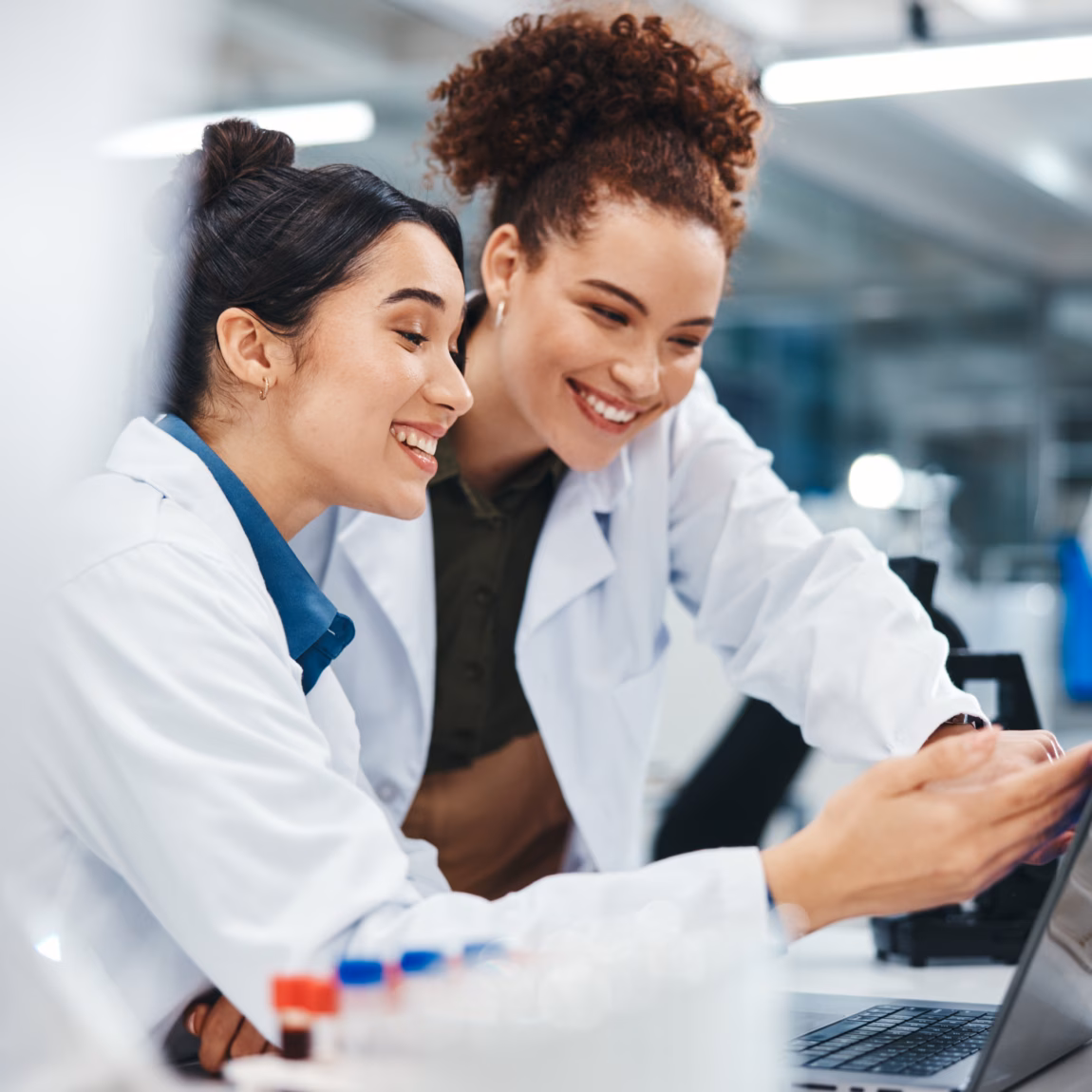 Zwei Frauen in Laborkitteln lächeln, während sie in einem Labor auf einen Laptop blicken; in der Nähe sind wissenschaftliche Geräte zu sehen.