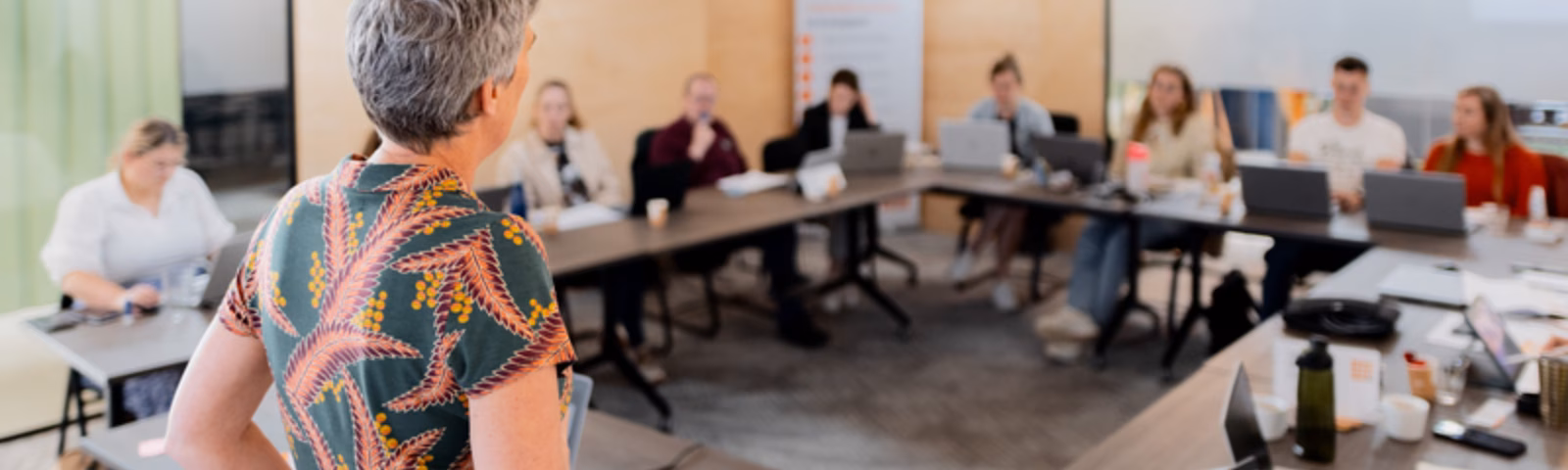 Participants attend a meeting or training session in a conference room while a speaker presents to the group.