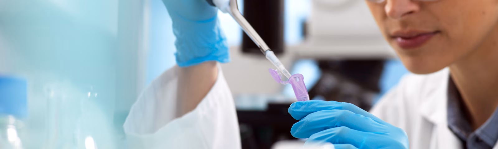 Een vrouwelijke onderzoeker met veiligheidsbril, blauwe handschoenen en witte labjas werkt geconcentreerd in het lab en pipetteert vloeistof in een reageerbuis.