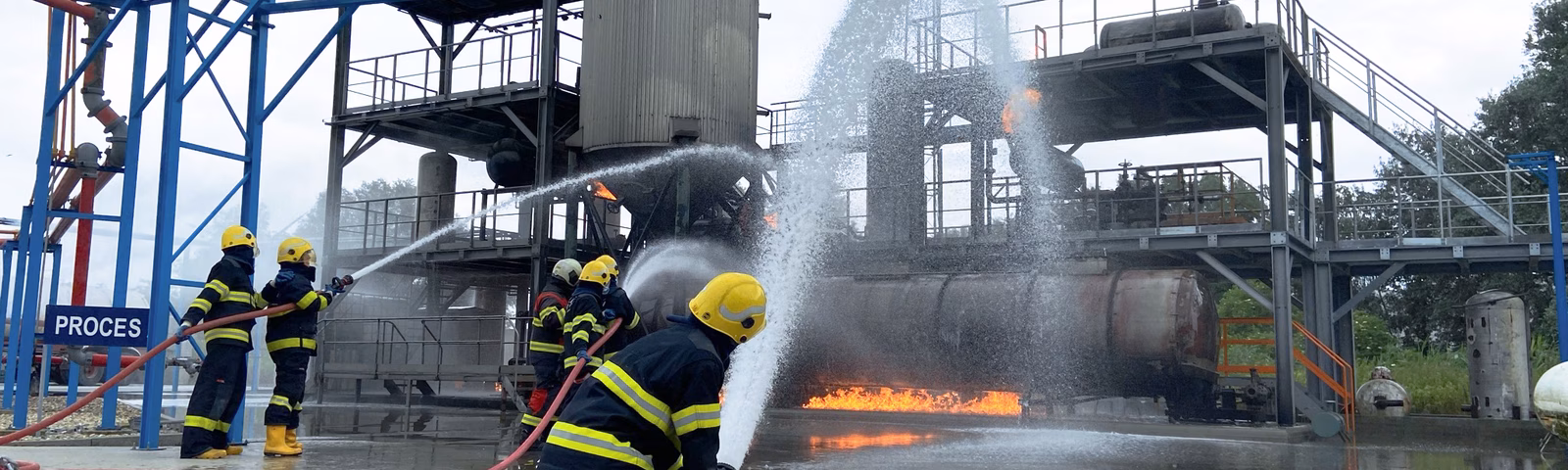 Firefighters are conducting a fire drill at an industrial facility with visible flames and water used for fire suppression.