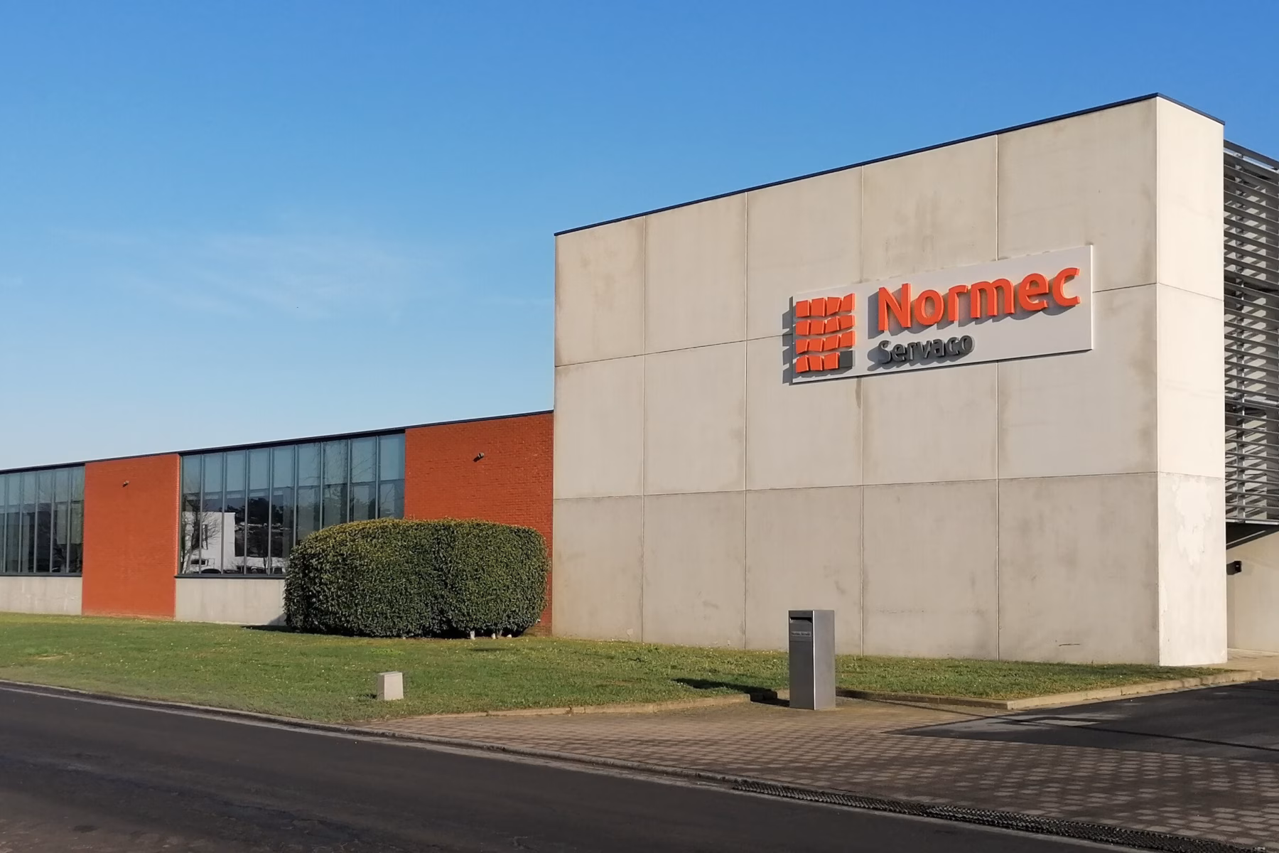Front view of the modern Normec Servaco building with white and dark grey façades, large windows and the Normec Servaco logo, photographed on a clear sunny day.