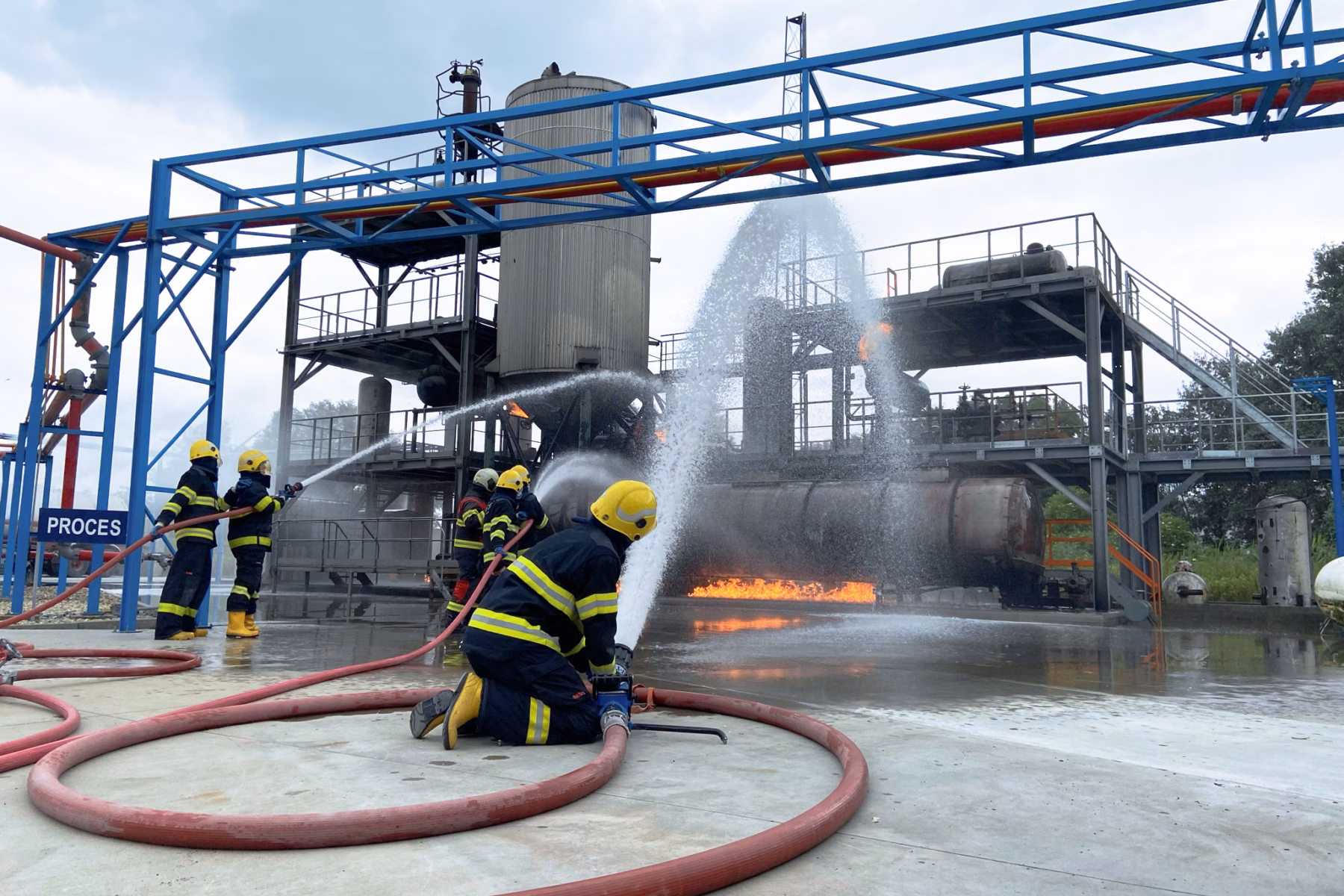 Firefighters in full firefighting gear are extinguishing an industrial fire using water.