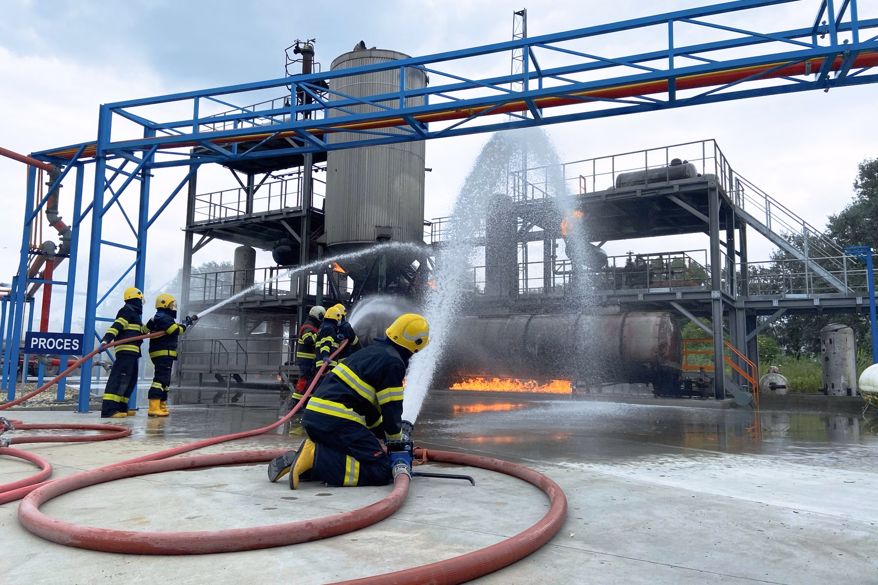 Firefighters are conducting a fire drill at an industrial facility with visible flames and water used for fire suppression.