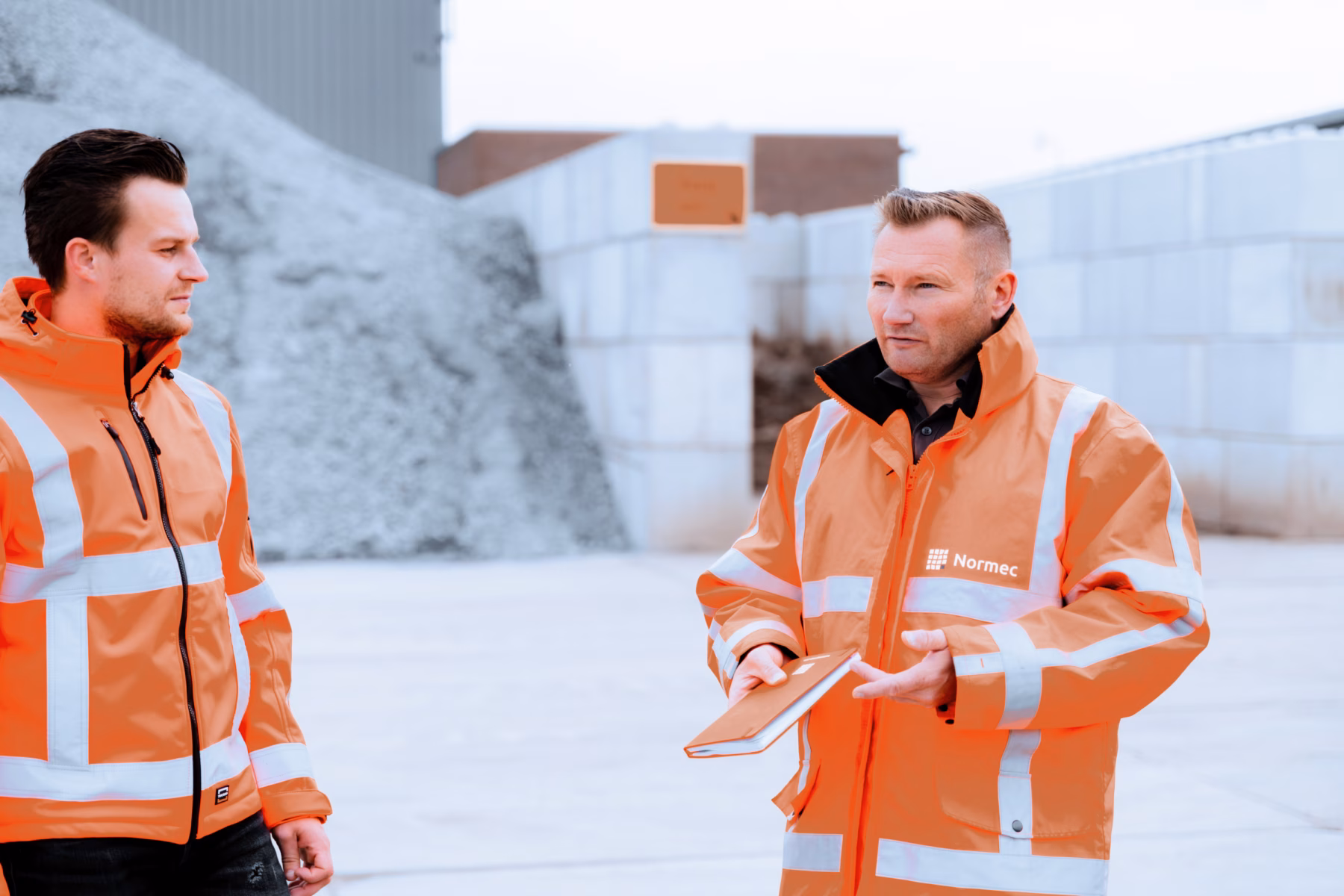 Men wearing orange coats are conversing on a construction site, with one man holding a notebook.