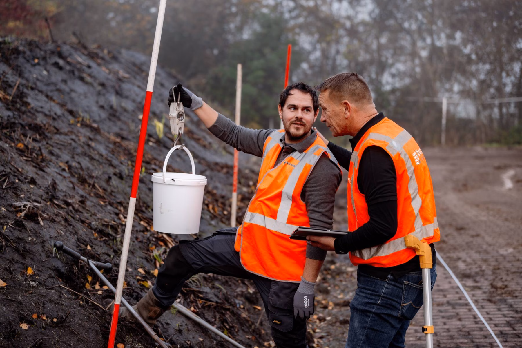 Men wearing orange reflective vests are inspecting a pile of soil, holding a bucket.